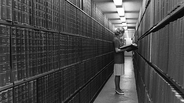 Vanha mustavalkoinen valokuva, jossa nainen seisoo kirja kädessä kirjavaraston hyllyjen välissä.