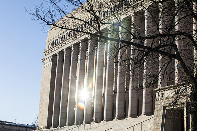 Eduskuntatalon julkisivua portaiden oikeasta laidasta kuvattuna. Edessä lehdetön puu, talon julkisivusta heijastuu auringon säde