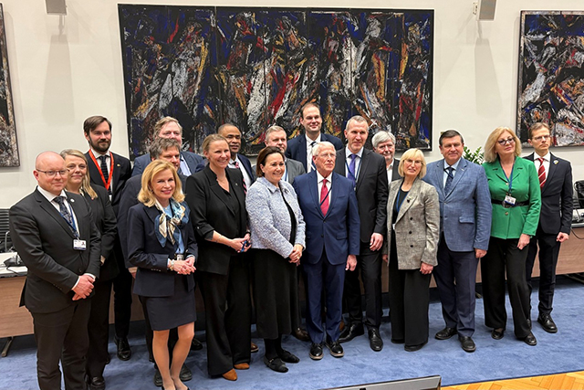 Noin 20 tapaamisen osallistujaa seisoo ryhmäkuvassa kokoushuoneessa taustallaan kookas tumma maalaus.