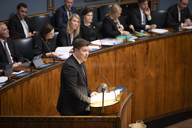 Kansanedustaja Tuomas Kettunen puhuu eduskunnan täysistuntosalin puhujankorokkeella. Takana istuu ministereitä ministeriaitiossa